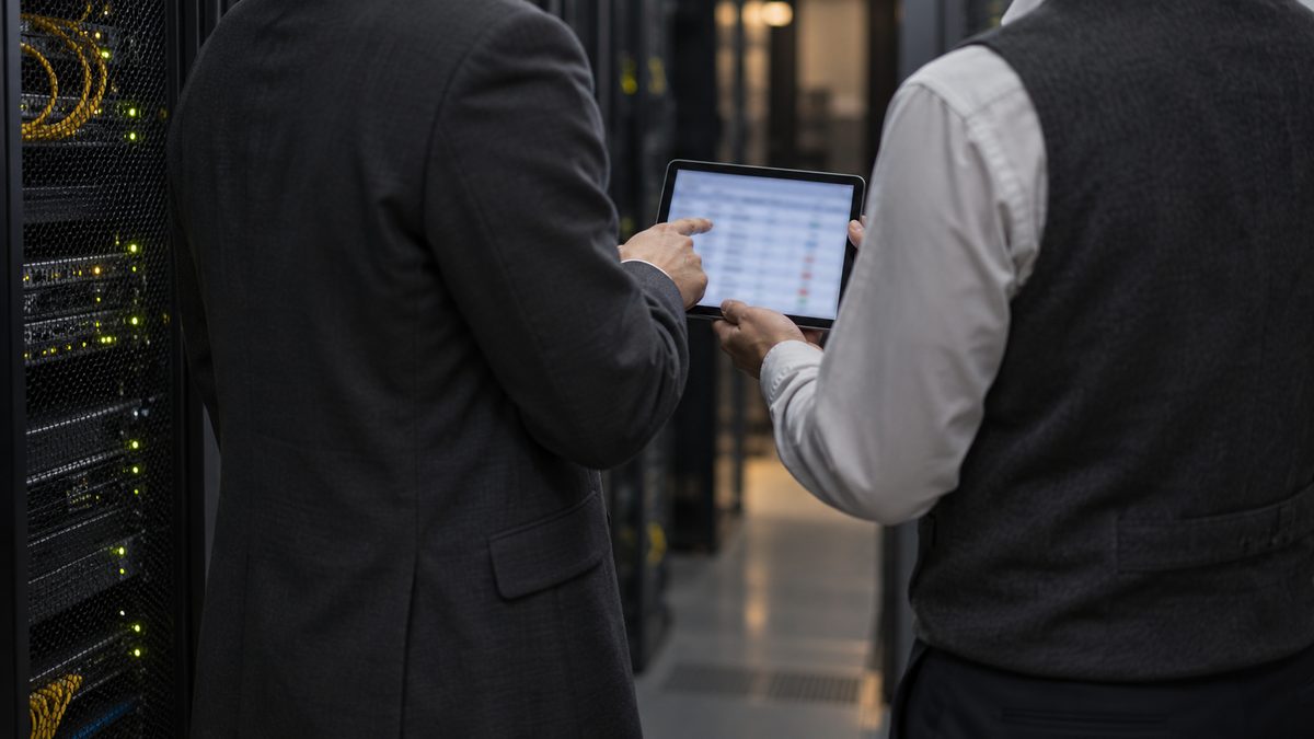 サーバー室のラックの前で、2人がタブレットの一覧表を指さしながら確認している後ろ姿