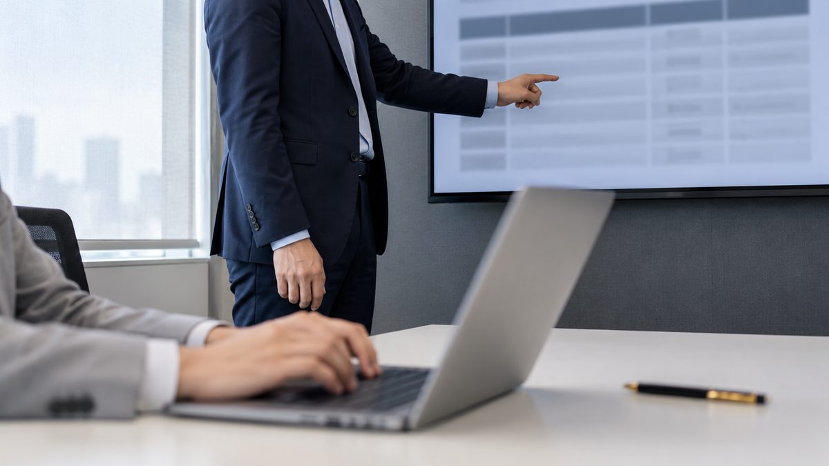 壁のモニターの表を示す外部技術者とノートPCに記録する社内担当