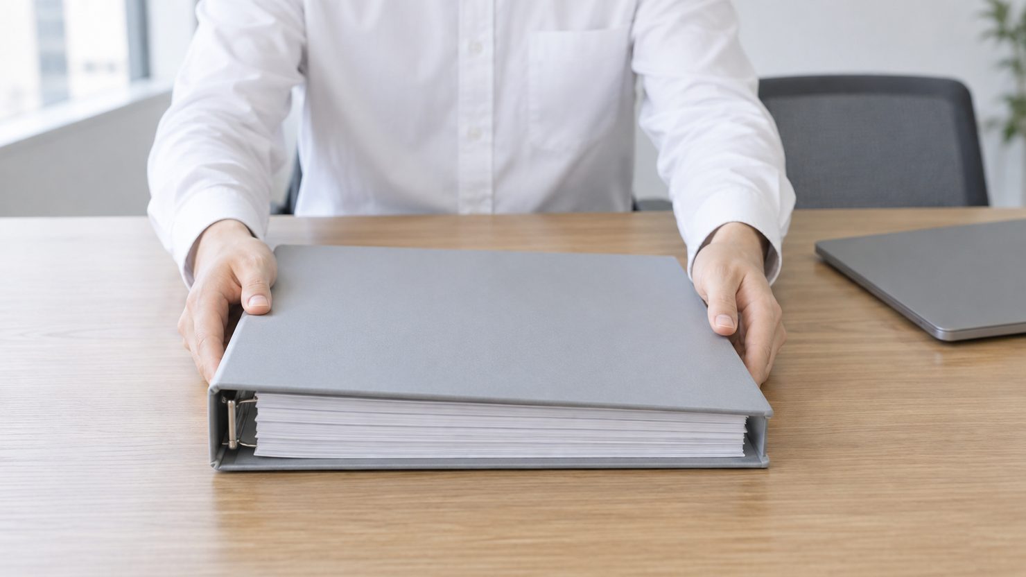 灰色の太いバインダーを机の中央に両手で置く、白いシャツの担当者。奥に閉じたノートPC
