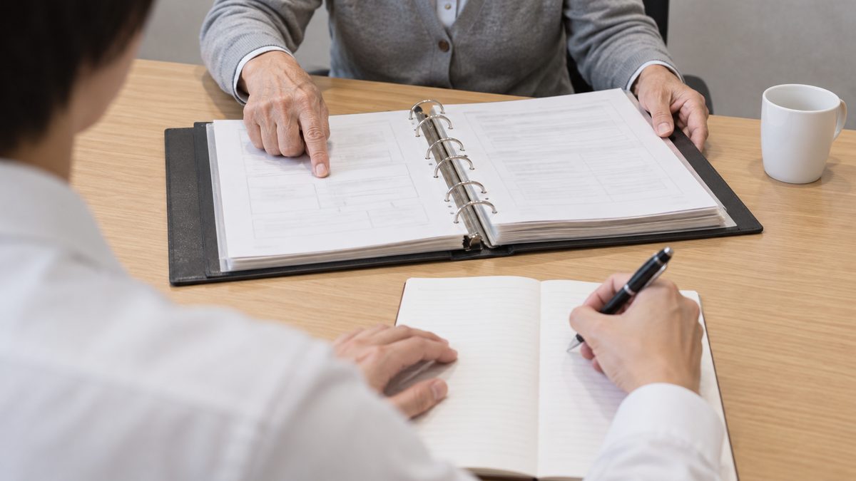 木目の机で年配の担当者が手順書のバインダーを開いて示し、手前の後任がノートに書き留めている