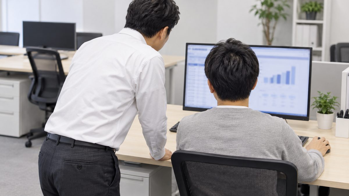 開発フロアで、座るエンジニアの横に白いシャツの上司が立ち、表と棒グラフの画面を一緒にのぞき込む