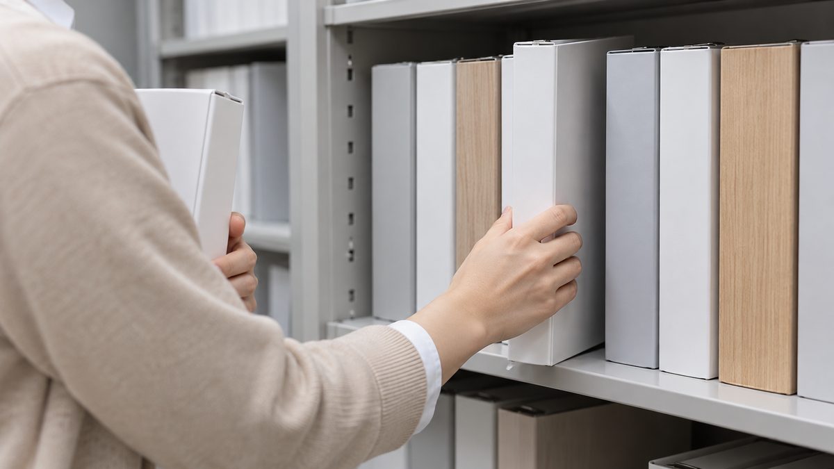 書類保管室のスチール棚で総務の担当者が無地の白いバインダーを列の隙間に差し込み、もう1冊を抱えている