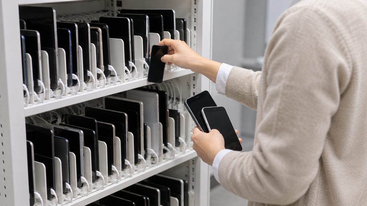 検証用のスマートフォンが並ぶ白い充電棚から担当者が1台を抜き取り、もう一方の手に2台を持っている