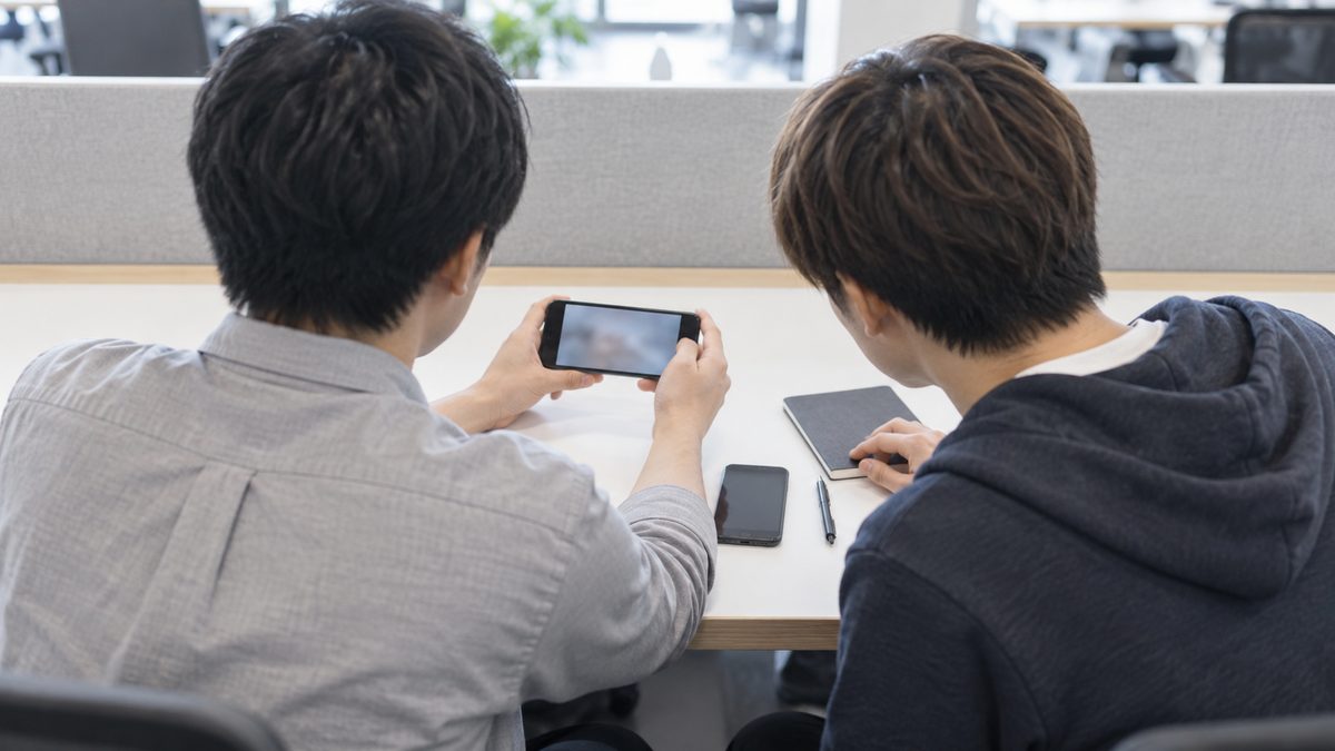 開発チームの席で2人が並んで座り、1人が横向きに持ったスマートフォンの画面をもう1人がのぞき込んでいる後ろ姿