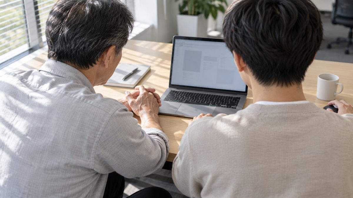 窓際の木目の机で50代のベテランと20代の若手のエンジニアが並んで座り、1台のノートパソコンを一緒に見ている後ろ姿