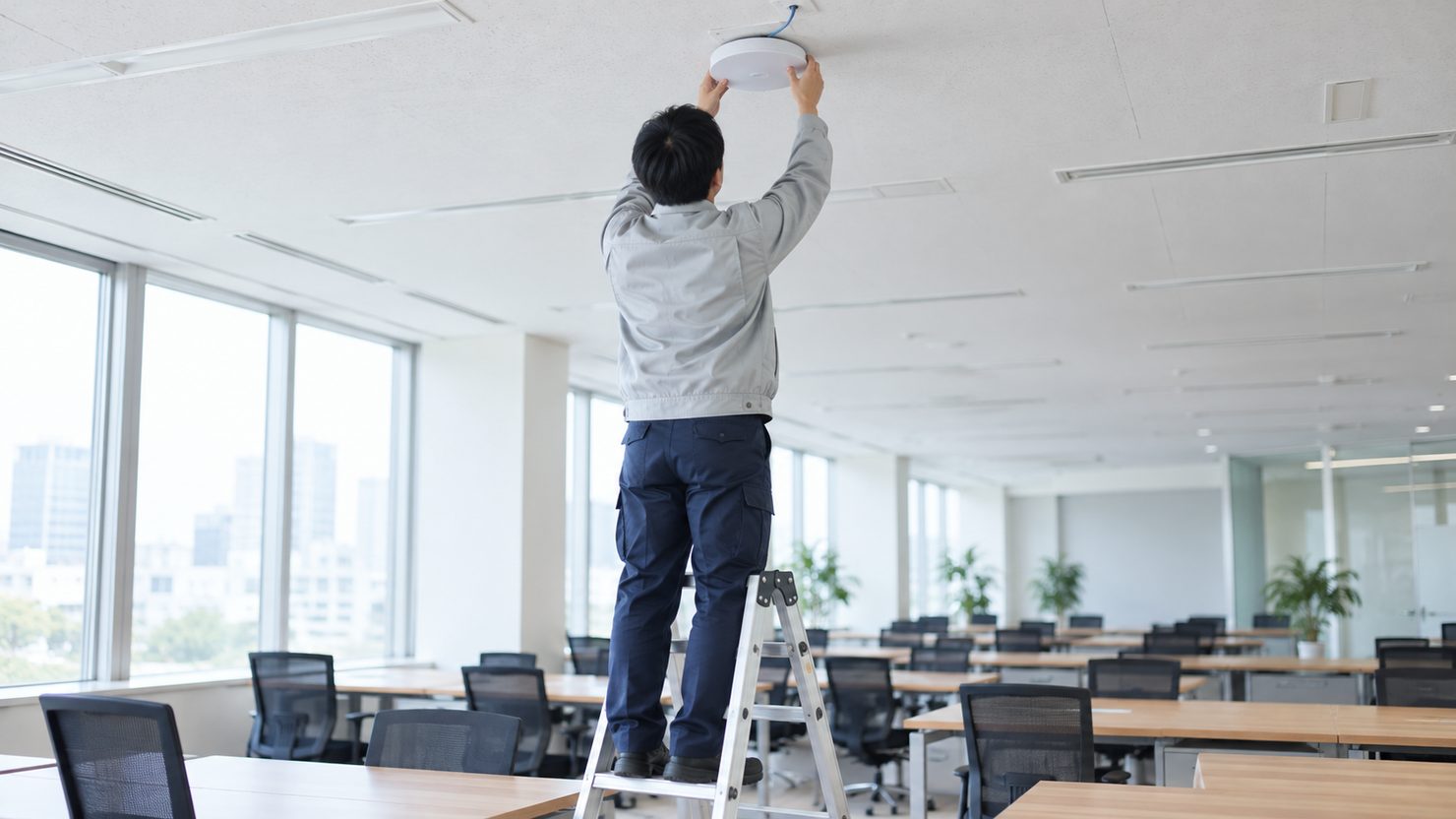 オフィスで脚立に乗った技術者が両手を上げて天井に白い円盤形の無線LAN機器を取り付ける後ろ姿