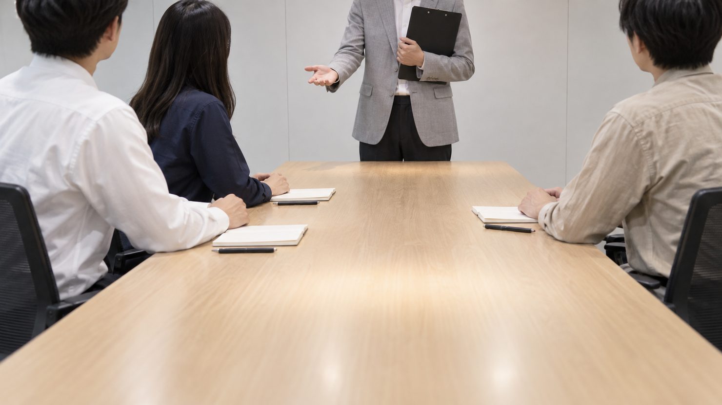 会議室の長机の端でクリップボードを抱えたPMOの担当者が手を差し出し、座った開発メンバー3人がその方を向いている