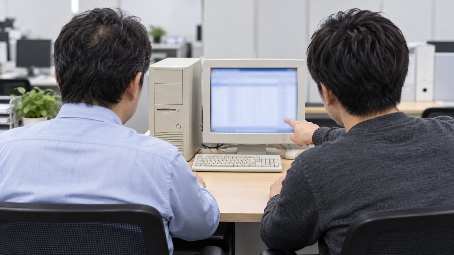 古いベージュ色のデスクトップ本体とモニターの前に並んで座り、画面を指して確かめる2人の後ろ姿