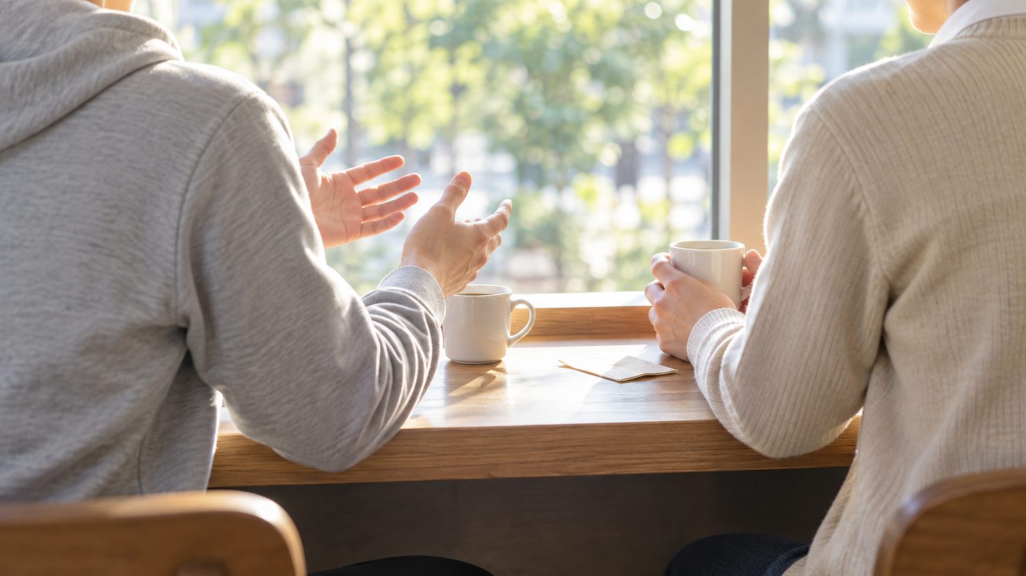カフェの窓際のカウンターで社員のエンジニアが手を広げて仕事の話をし、隣の元同僚がカップを持って聞いている