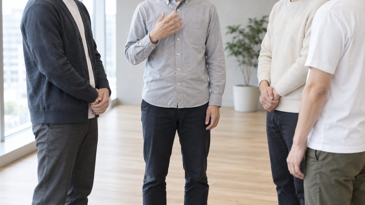 窓際で立ったまま輪になる朝会で、新しく加わった技術者が胸に手を当てて話し、3人が体を向けて聞いている
