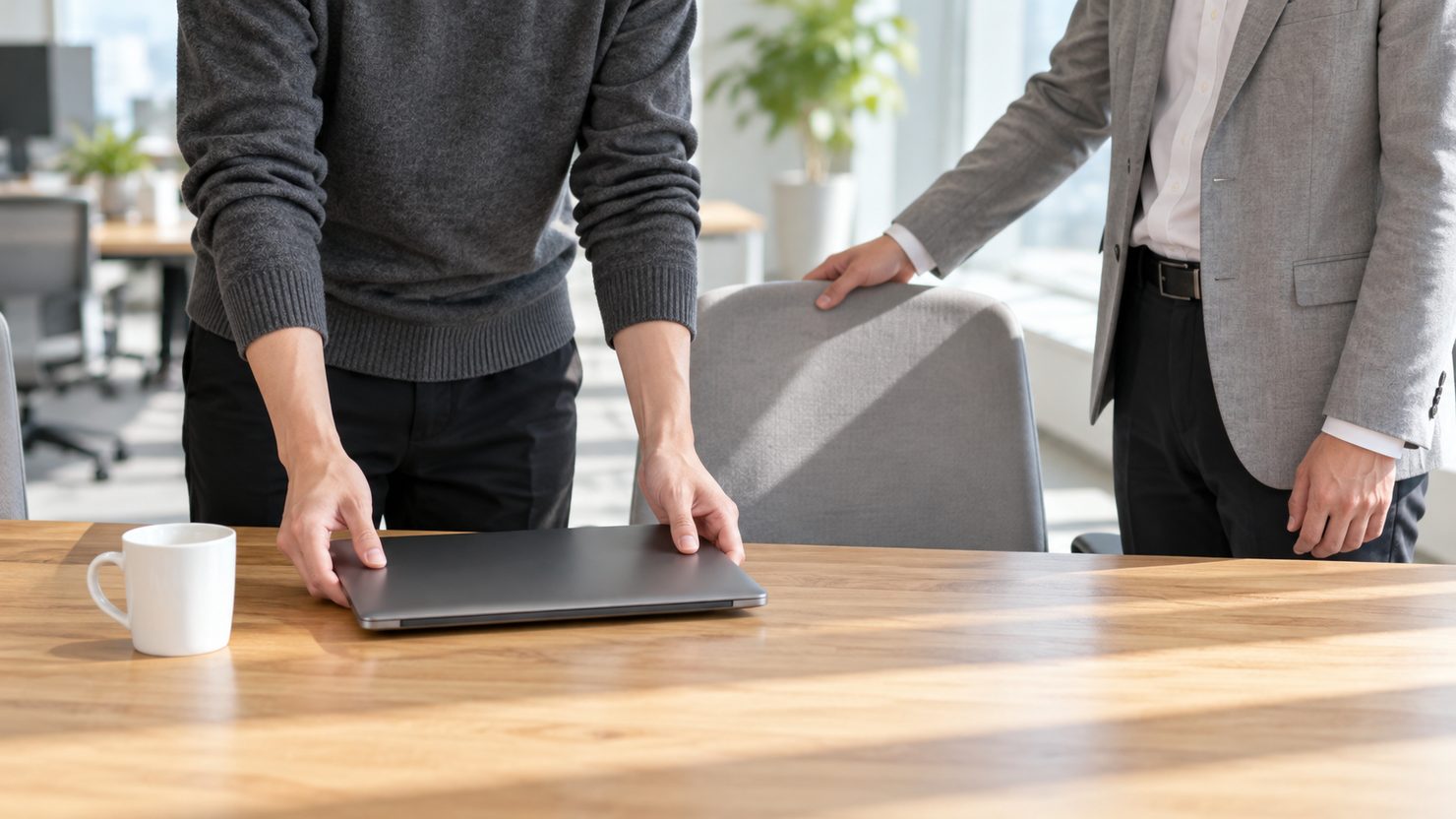 開発チームの木目の机に閉じたノートパソコンを置くエンジニアと、椅子の背に手を添えて席を示す開発マネージャー